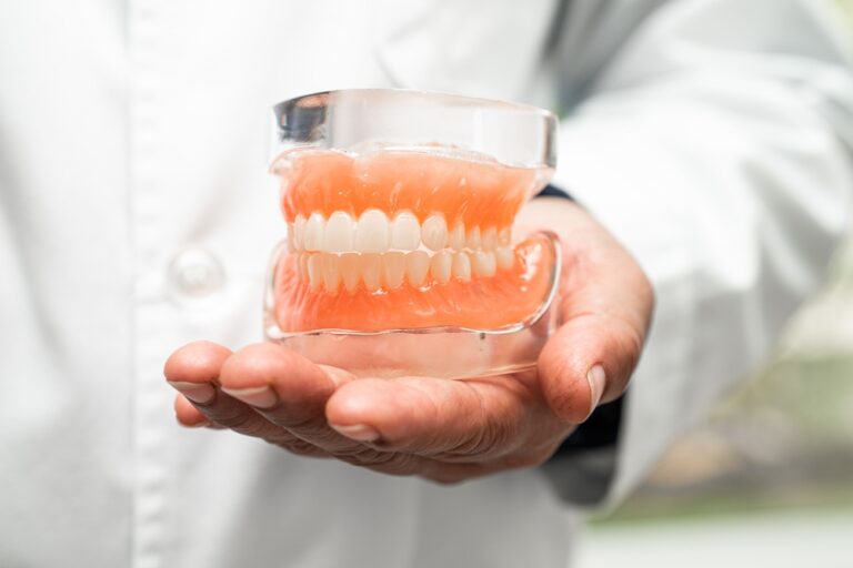 A person in a white coat holds a realistic dental model of upper and lower gums with white teeth. The background is blurred, highlighting the hand and dentures.