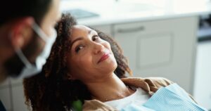 A woman with curly hair smiles while sitting in a dental chair. A dentist wearing a face mask looks at her in a bright, modern office.