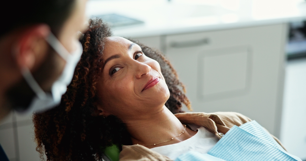 A woman with curly hair smiles while sitting in a dental chair. A dentist wearing a face mask looks at her in a bright, modern office.