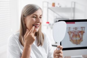 A middle-aged woman with grey hair smiles while pointing to her teeth in a hand mirror. She wears a white shirt in a medical office, with a computer monitor showing a 3D dental scan of a jaw in the background.