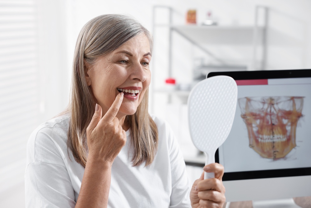 A middle-aged woman with grey hair smiles while pointing to her teeth in a hand mirror. She wears a white shirt in a medical office, with a computer monitor showing a 3D dental scan of a jaw in the background.